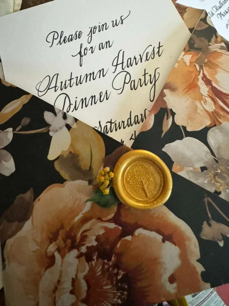 Invitation to an autumn harvest dinner party with elegant script, floral-patterned wrapping paper, and a gold wax seal with a tree emblem.