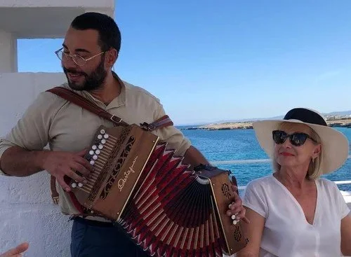 A man playing an accordion on a boat or seaside with an older woman wearing sunglasses and a wide hat, with the ocean and sky in the background.