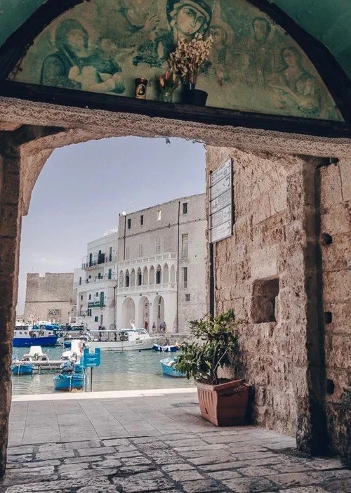 View of a harbor with boats, framed by a stone archway with religious iconography and plants inside a building