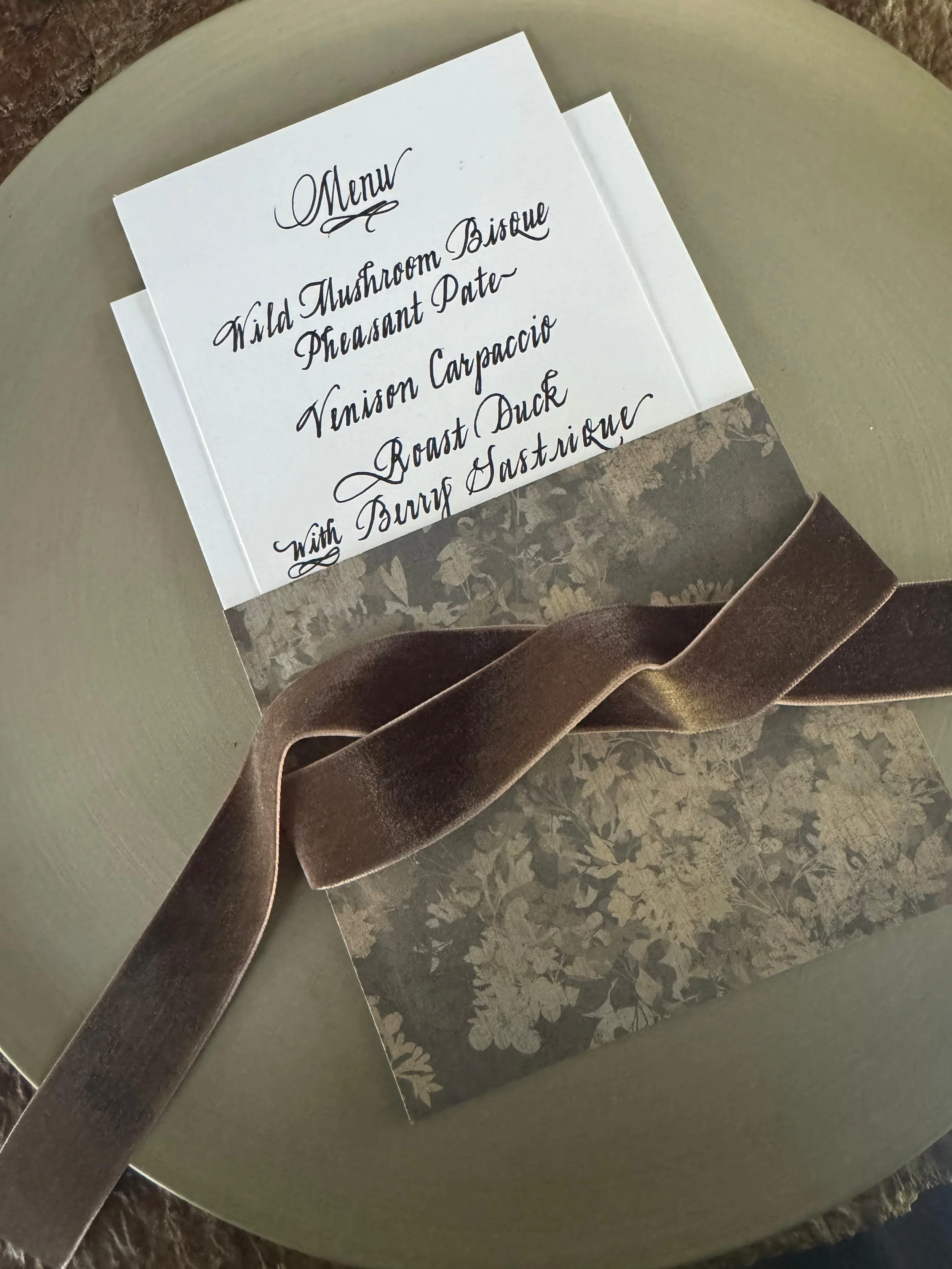 A menu card placed on a green plate, with a chocolate-brown ribbon and a floral-patterned napkin underneath.