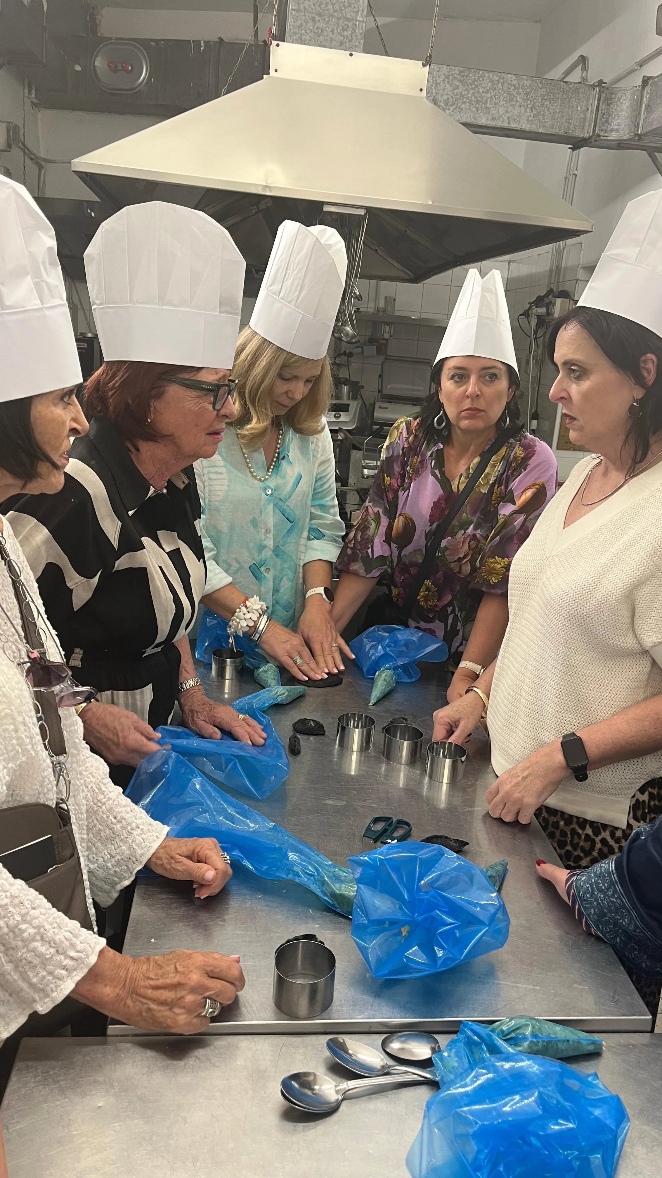 A group of women wearing chef hats gathered around a stainless steel table in a kitchen or food processing area, discussing or demonstrating something with blue plastic bags and metal cookie cutters on the table.