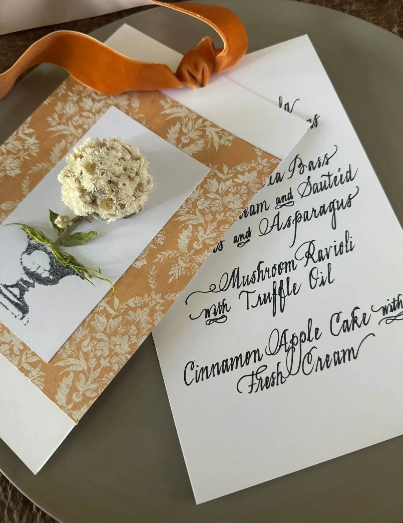 A white flower boutonniere with small white flowers and green leaves tied with green string, placed on a decorative brown and white floral-patterned paper. A paper with handwritten calligraphy describing a menu or event, including dishes like mushroo
