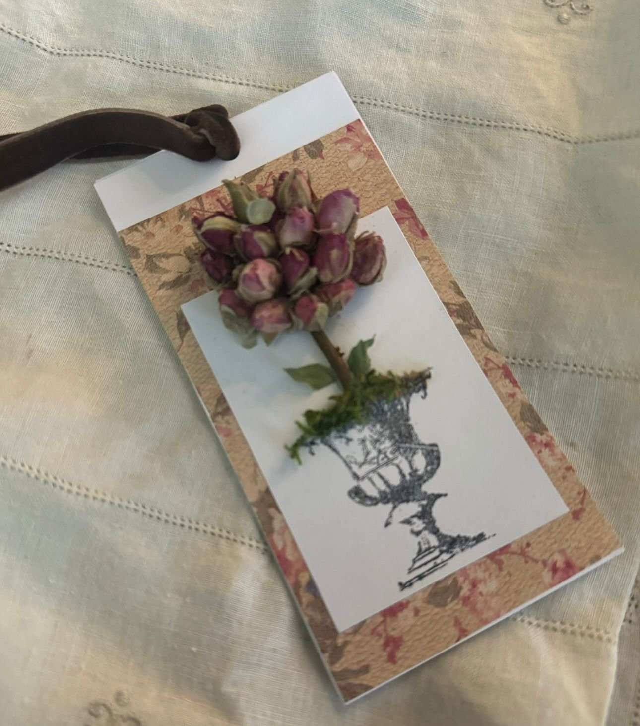 A dried pink rosebud on a decorative paper with a black and white illustration of a vintage lamp, set on a cream-colored fabric surface.