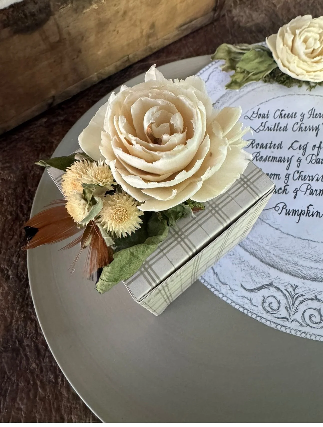 A white rose adorns a decorative gift box with smaller dried flowers and a feather. The box is on a gray plate next to a cloth with a handwritten menu.