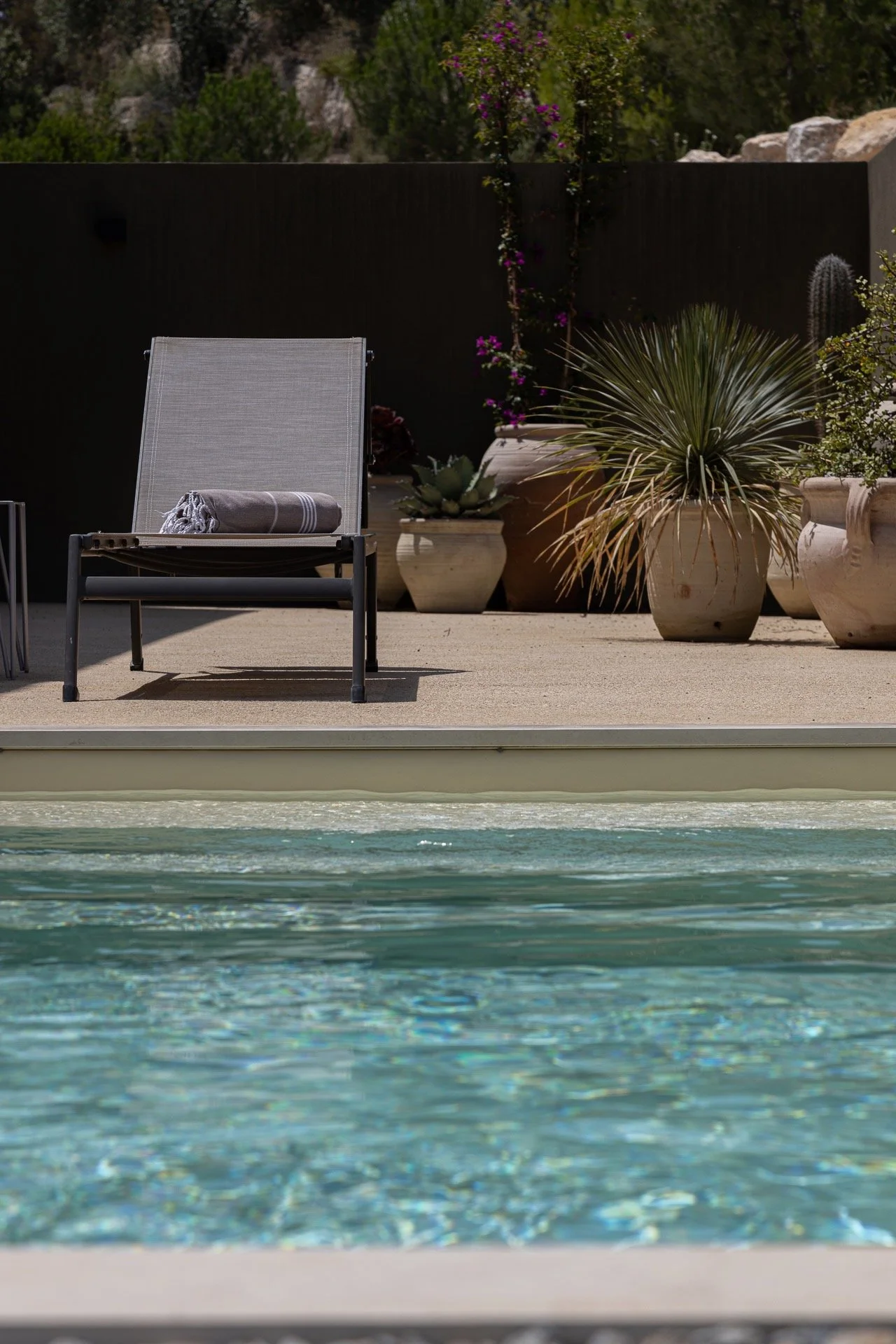 Patio scene with pool in the foreground, sitting area with a lounge chair and rolled towel, and large potted desert plants in the background.