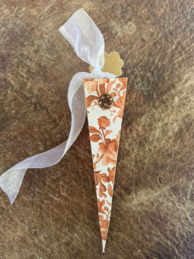 Decorative paper cone with floral pattern, adorned with a black and gold swirl ornament, a white ribbon, and a small beige leaf tag, placed on a brown textured surface.
