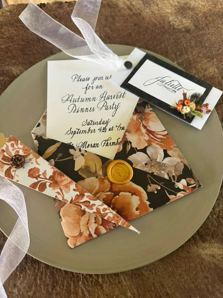 A floral autumn-themed wedding invitation set includes an envelope sealed with a gold wax stamp, a place card with a floral design, and an RSVP card, all arranged on a light-colored plate with a ribbon.