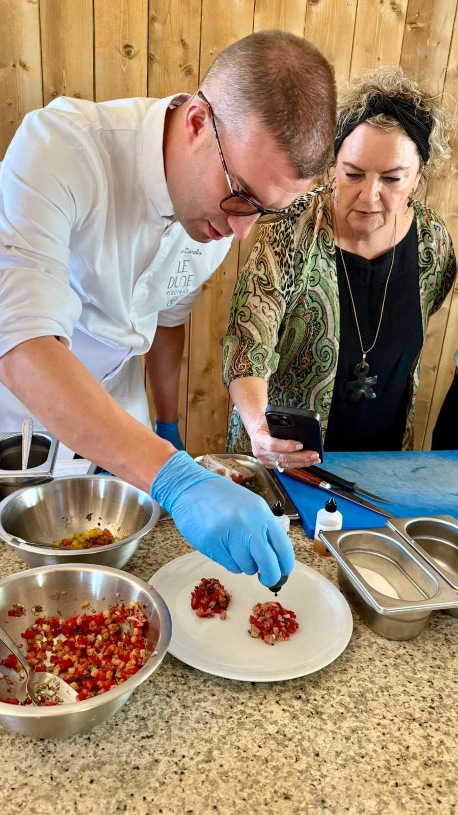Two women and a man preparing food in a kitchen with wooden walls. The man is wearing a white coat, blue gloves, and glasses, and is garnishing dishes with a squeeze bottle, while the woman next to him, holding a smartphone, observes closely. Various bowls of chopped vegetables and ingredients are on the counter.