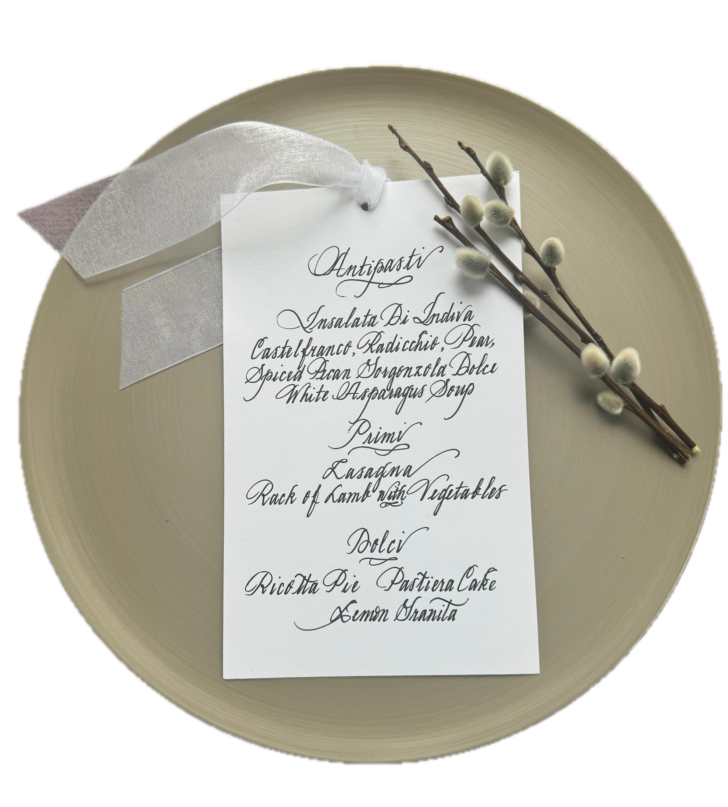 A circular beige plate with a white handwritten menu card attached with a white ribbon, and sprigs of pussy willow branches placed over the card.