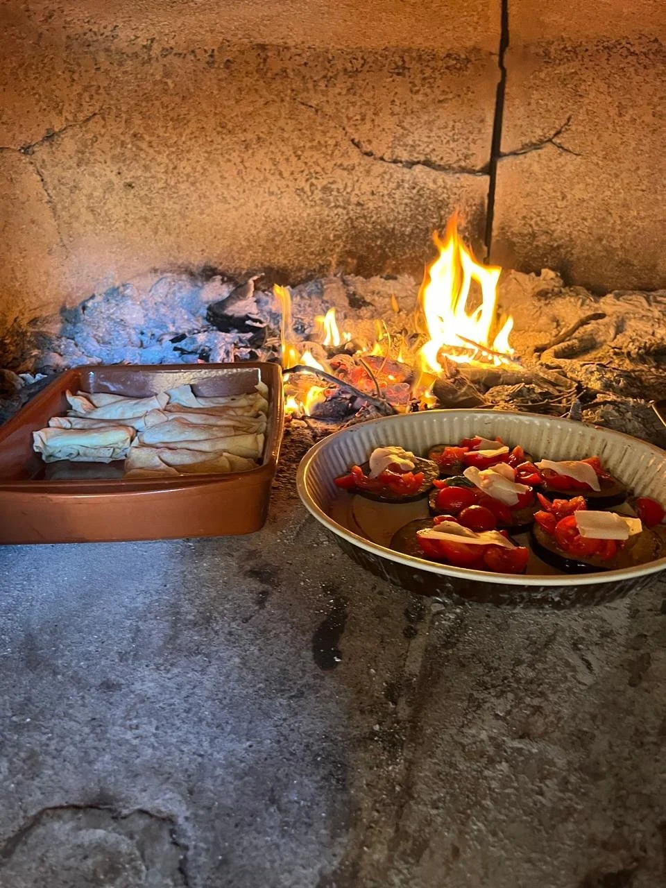 An indoor or outdoor pizza oven with an open flame, preparing to cook items on a baking tray and a plate of sliced tomatoes topped with cheese.