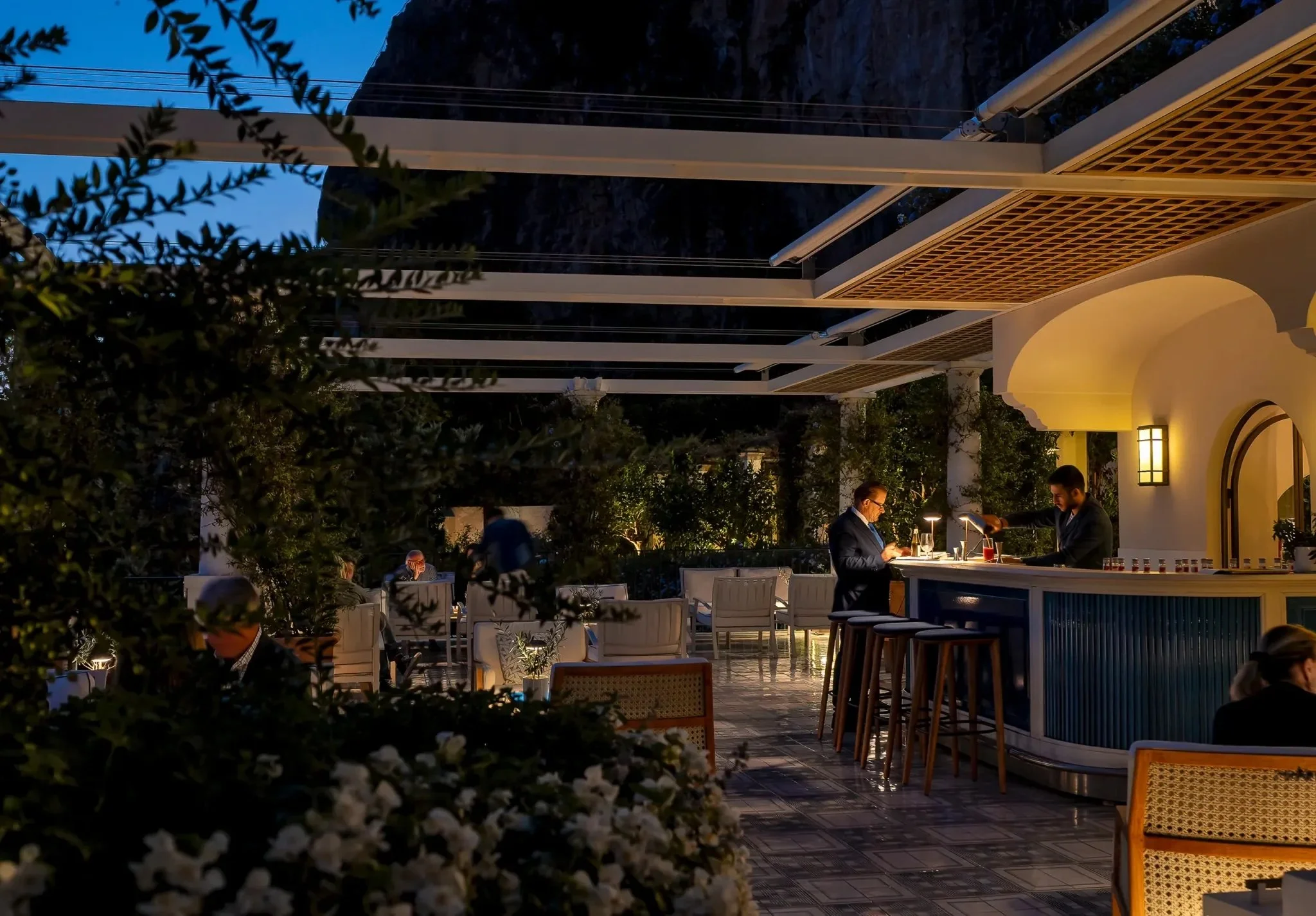 An outdoor bar area at dusk with a bartender preparing drinks, patrons seated at tables, and lush greenery illuminated by warm lighting.