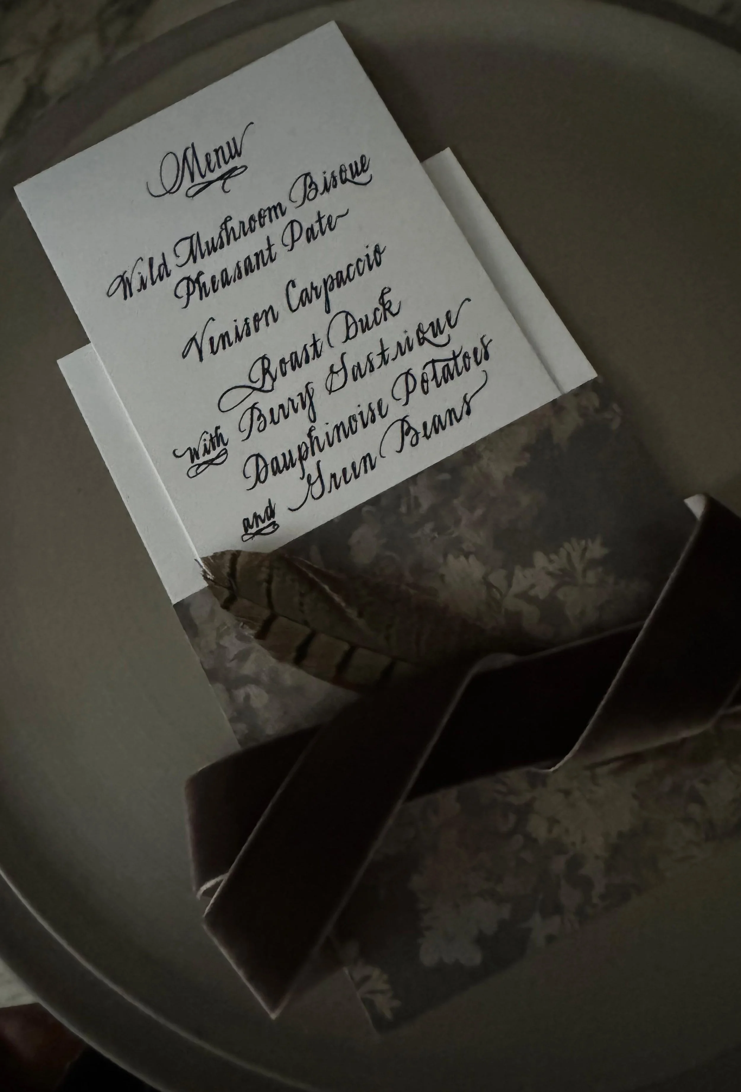 A handwritten menu listing dishes such as wild mushroom bisque, pheasant pate, venison carpaccio, roast duck, and dauphinoise potatoes, placed on top of a round container.