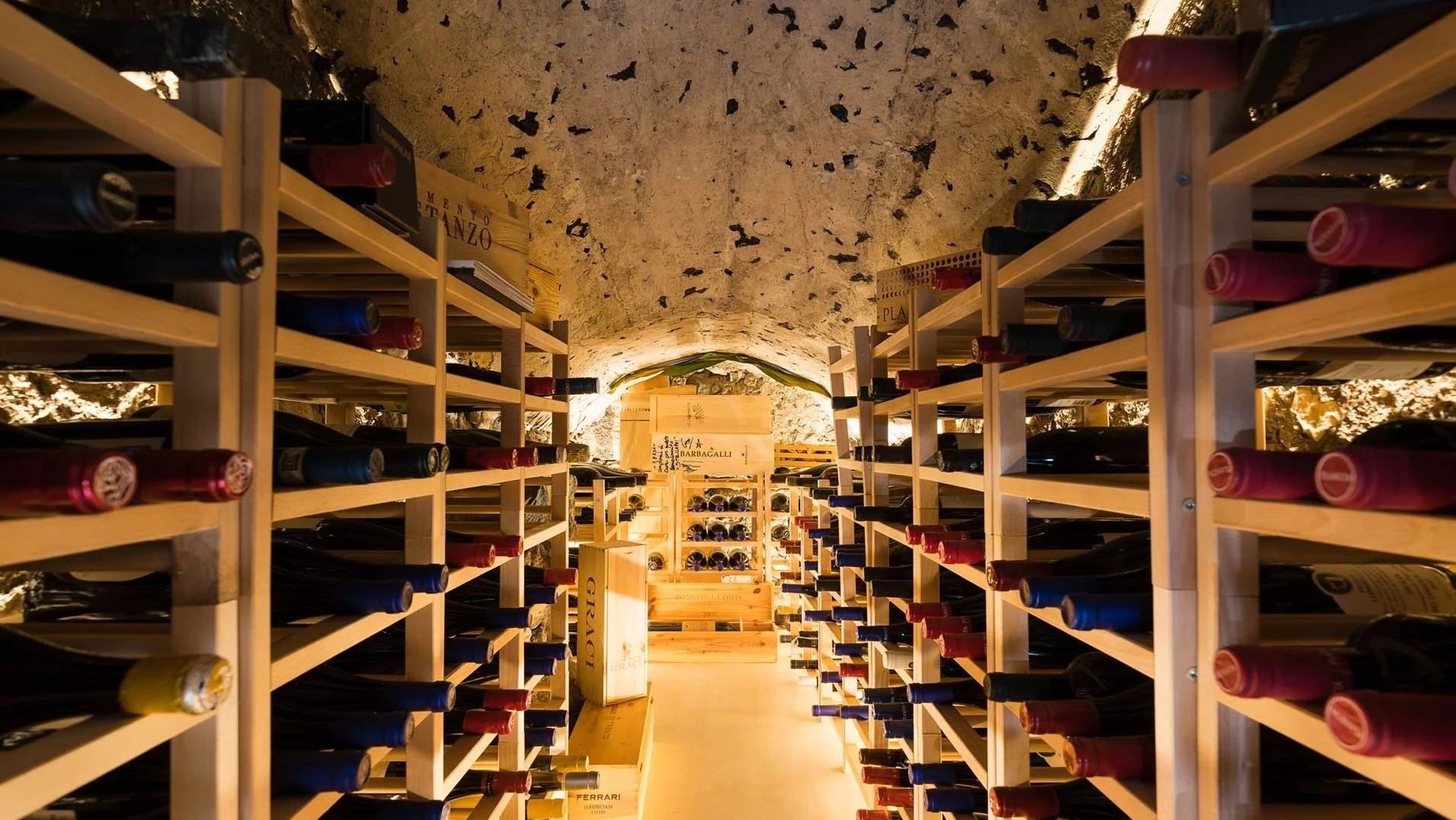 Wine bottles stored on wooden racks in a tunnel-like wine cellar with a stone ceiling and warm lighting.