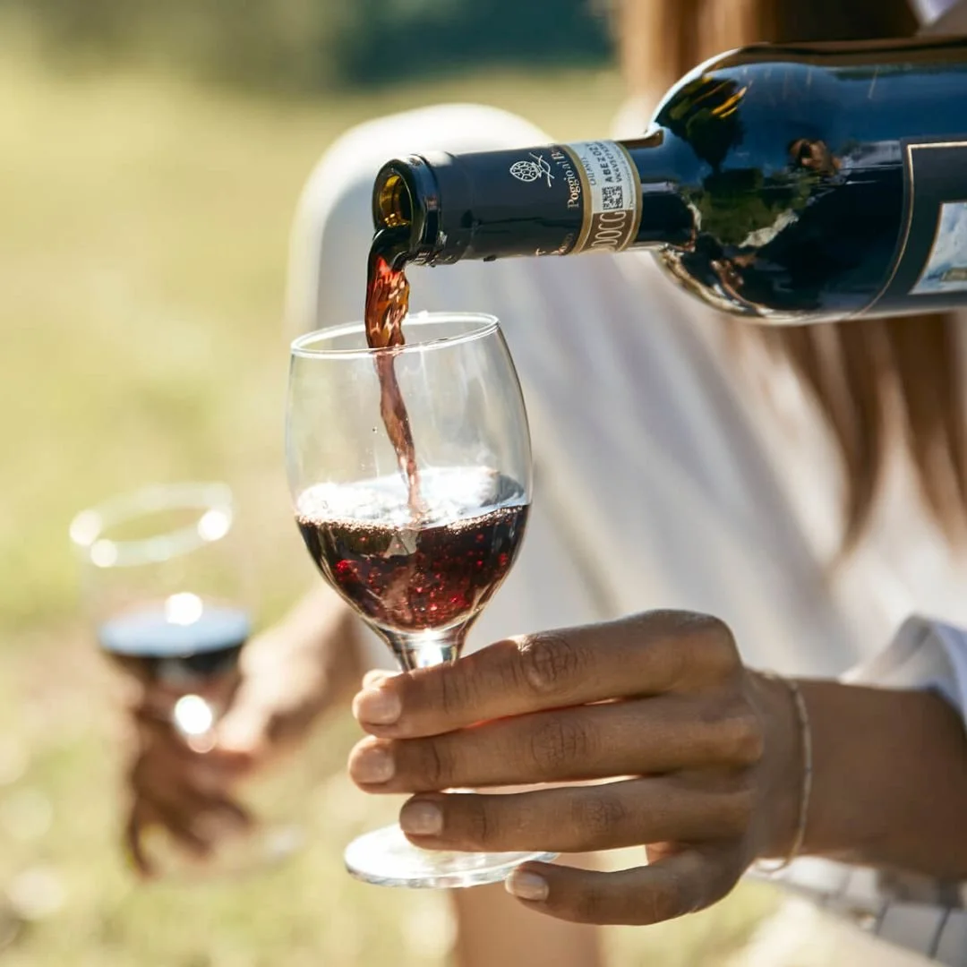 A person pouring red wine into a wine glass outdoors with another person holding a glass in the background.