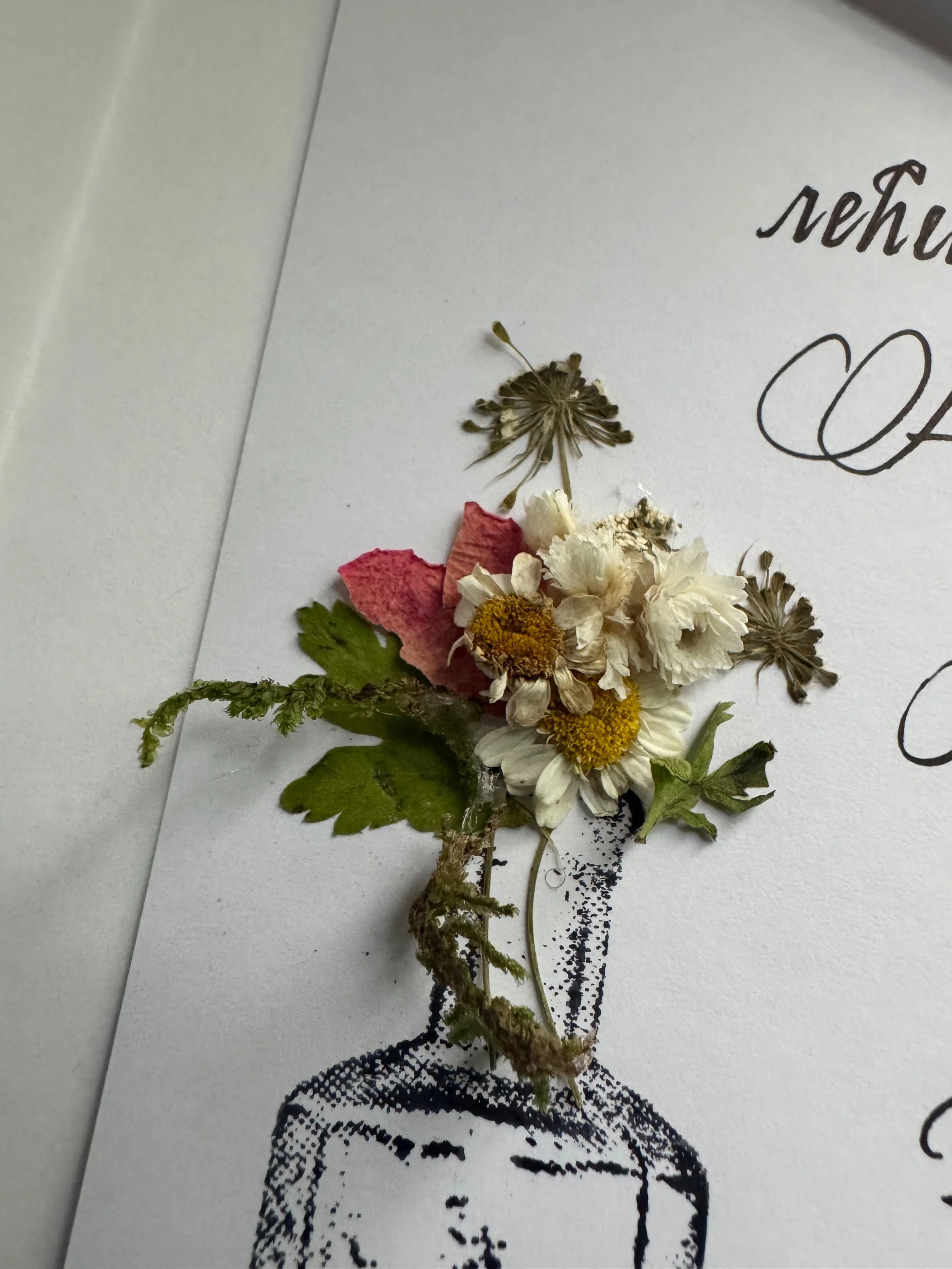 Pressed and dried flowers arranged on a white sheet with decorative black ink pattern and cursive writing.