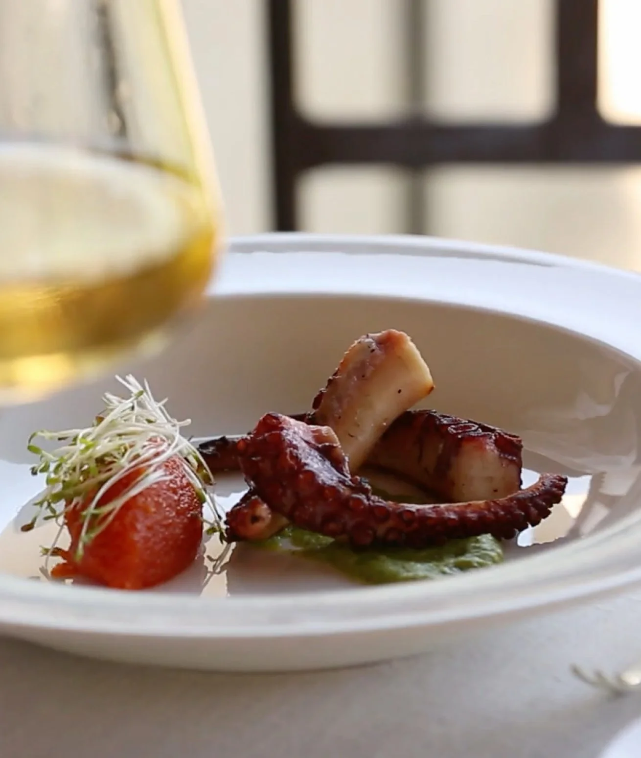A white plate with grilled octopus tentacle, roasted cherry tomato with sprouts, and green sauce, with a glass of white wine in the background.