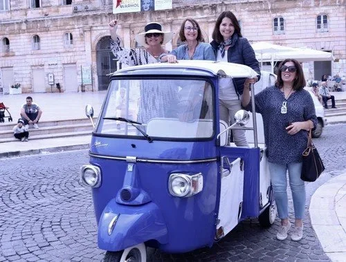 Four women standing around a blue auto rickshaw in a historic square, with a stone building and steps in the background.