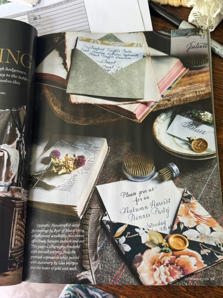 Open magazine showing an autumn-themed invitation for a dinner party, alongside calligraphy supplies, a small floral arrangement, and decorative seals on stationery.
