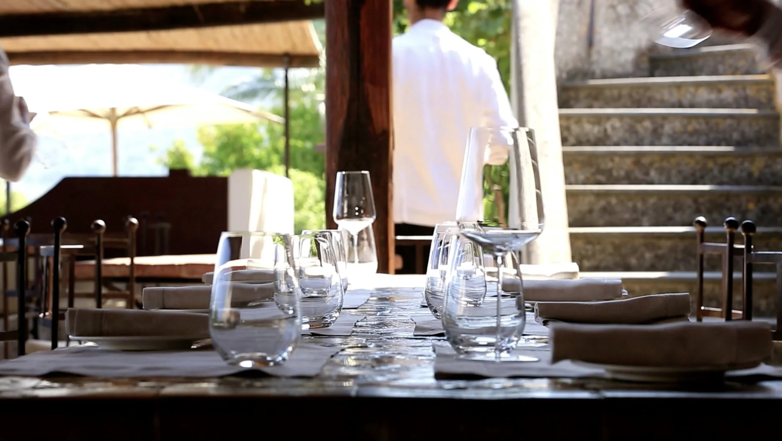A table set with multiple wine glasses and napkins, in an outdoor dining area with a view of greenery and stairs in the background.
