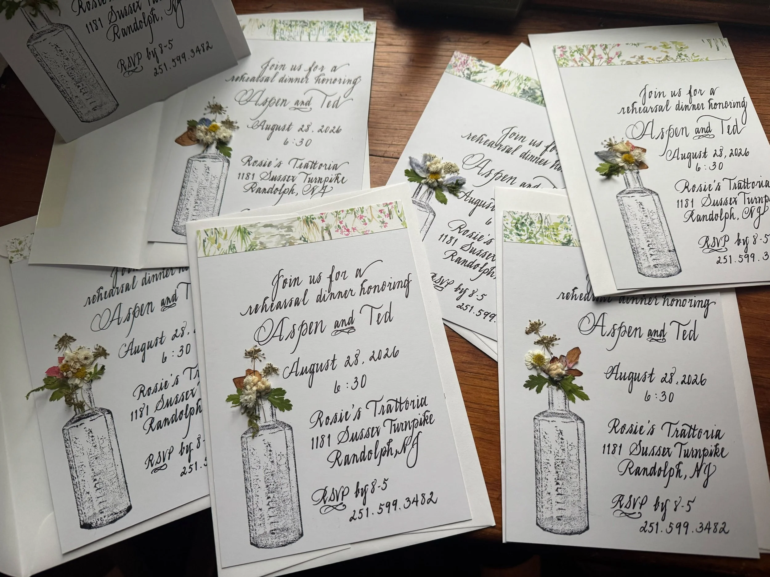 Multiple wedding invitations featuring a vintage ink drawing of a bottle with flowers, handwritten styled text with event details, and decorative floral tape accents.