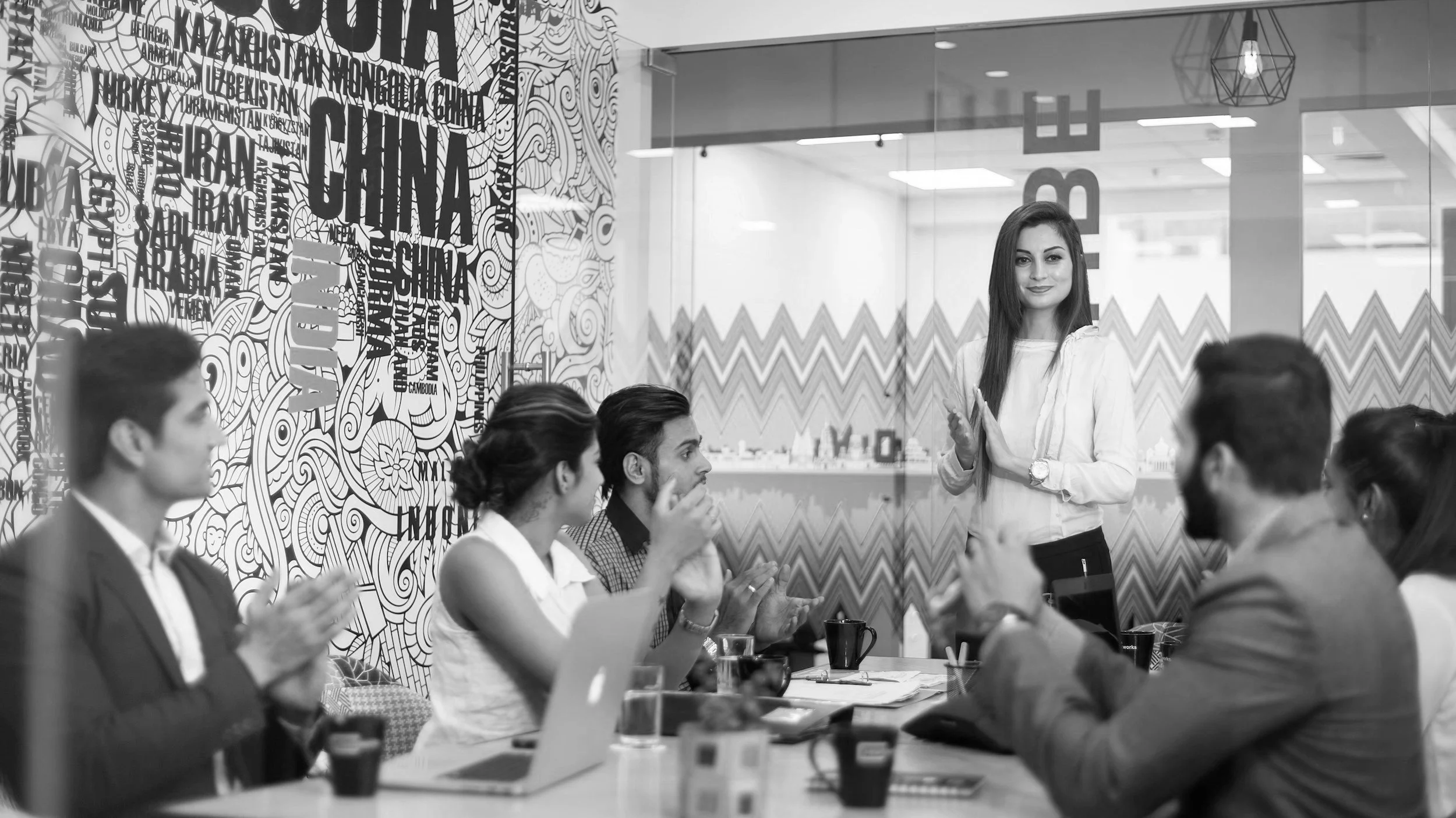 A woman standing in front of a group of people in a meeting room, gesturing with her hands as she speaks. The attendees are sitting around a table with laptops, notebooks, and cups, and are listening attentively.