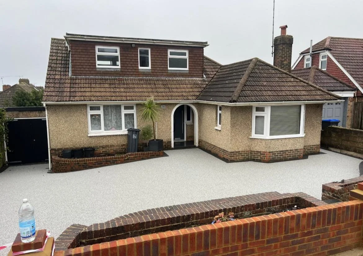 A house with a gravel driveway and a brick wall in the foreground. The house has a two-story section and a single-story section with a rounded entrance door, a small front garden bed, and multiple windows.