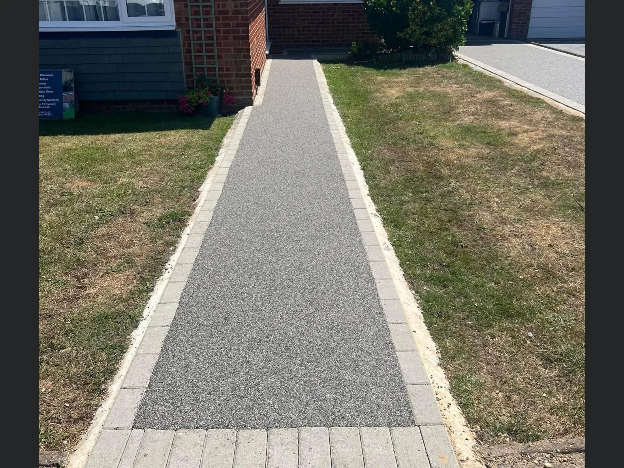 A concrete sidewalk bordered by gray bricks on both sides, leading towards a house with a blue and brick facade. There are patches of grass on each side of the sidewalk, with the grass on the right appearing patchy and dry, while the grass on the lef
