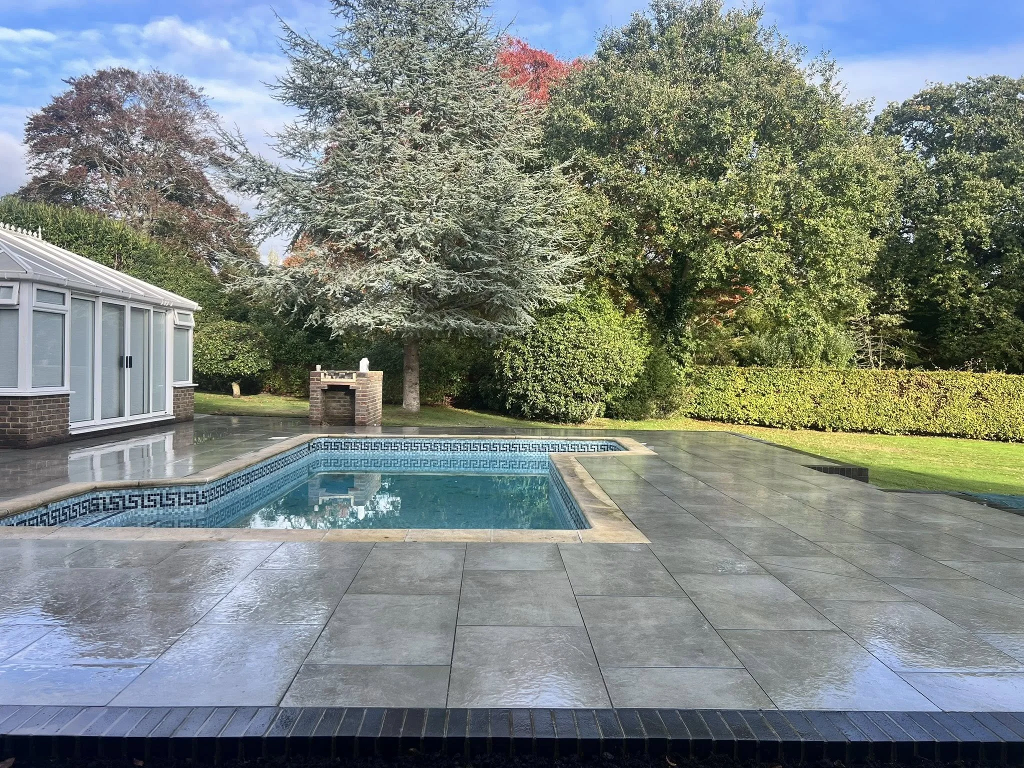 Empty backyard with a swimming pool, surrounded by trees and bushes, and a small brick barbecue grill, with wet stone patio surface.