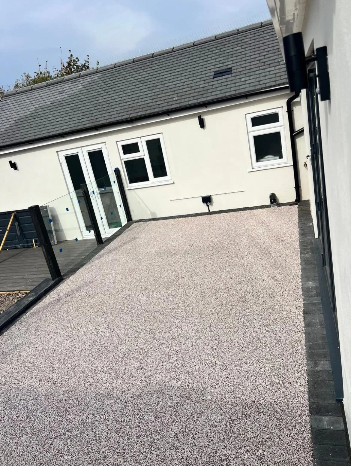 Backyard deck with a textured surface, glass railing, and a white house with multiple windows and a sliding door.