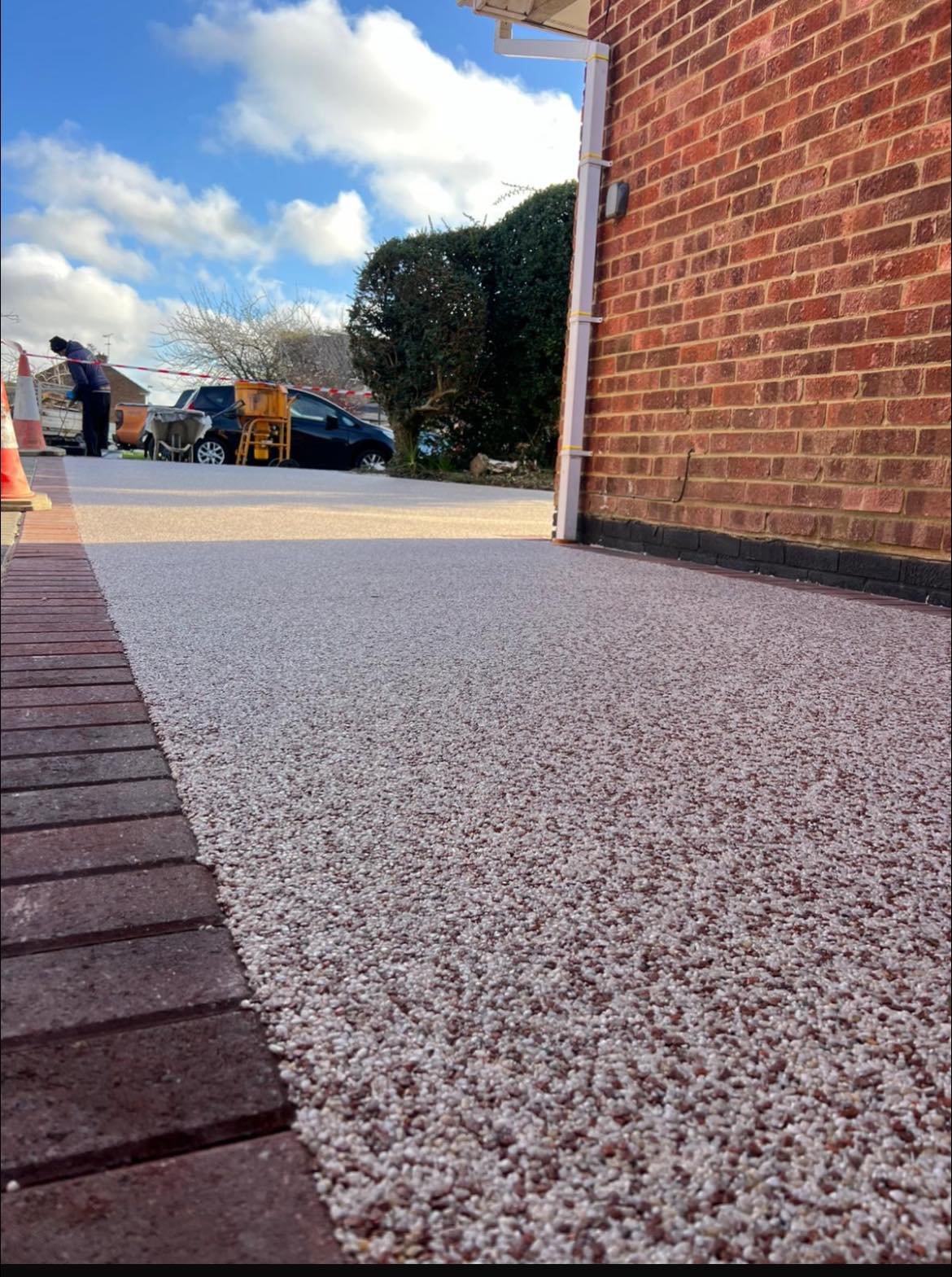Close-up of a freshly installed pebble driveway next to a brick house, with a view of the street and cars in the background under a partly cloudy sky.