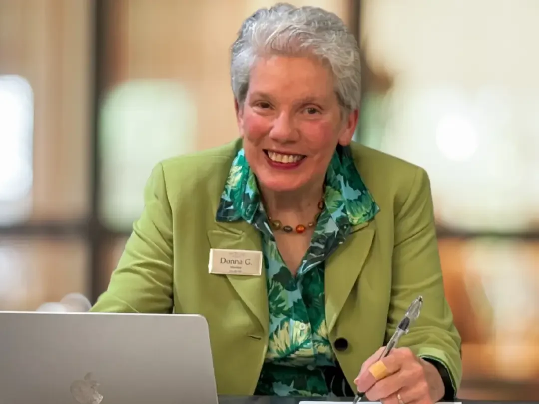 Donna R. Gore sitting at a desk with a green jacket and print blouse. Sitting in front of a laptop with a pen in her hand.