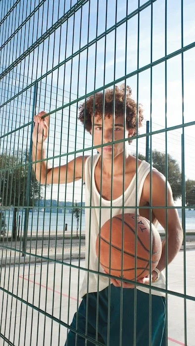 A teen boy holding a basketball behind a fence.