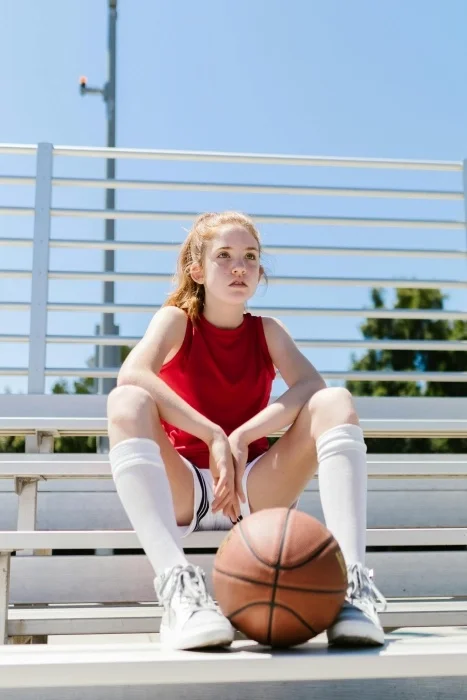 A teenage basketball player in a red tank top and tall white socks, sitting on metal bleachers with a basketball between her feet.