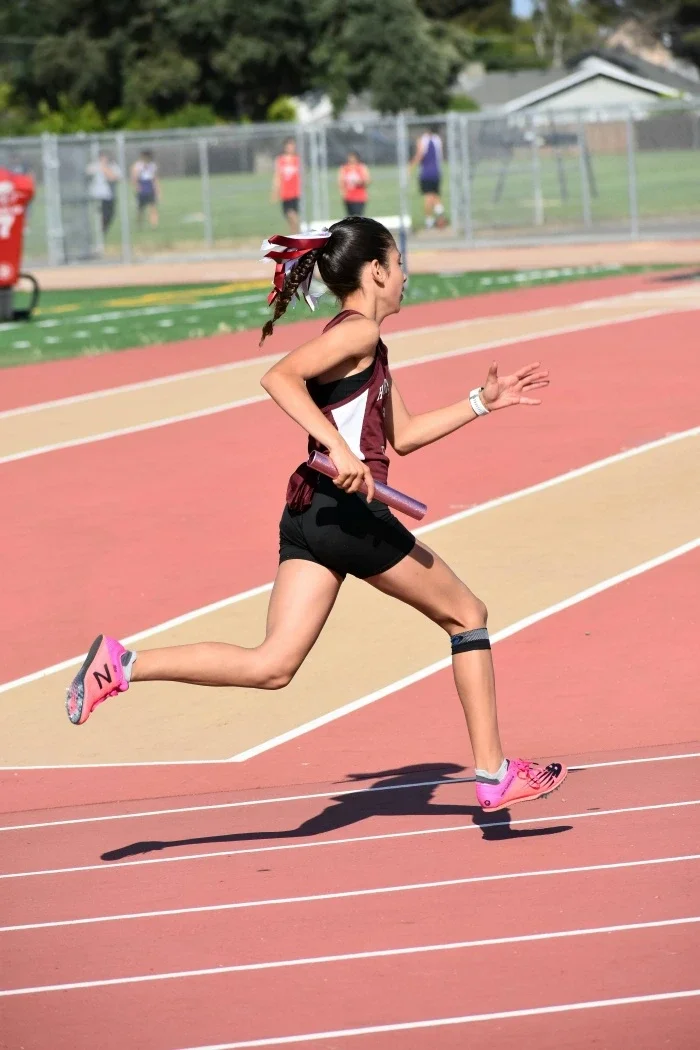 A high school track runner running on the track in red and white, holding a baton.