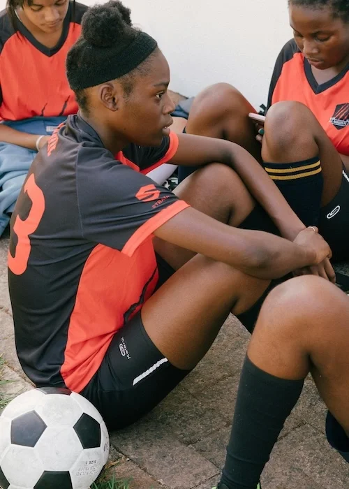 A girl soccer player sitting with her team on the ground. She looks nervous.