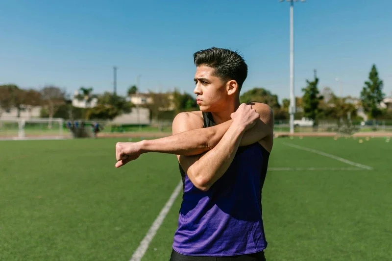 A teen boy stretching his arm on an outdoor sports field.