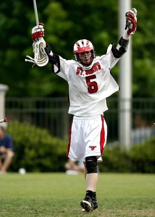 A high school lacrosse player in his red and white uniform, holding his crosse up into the air.
