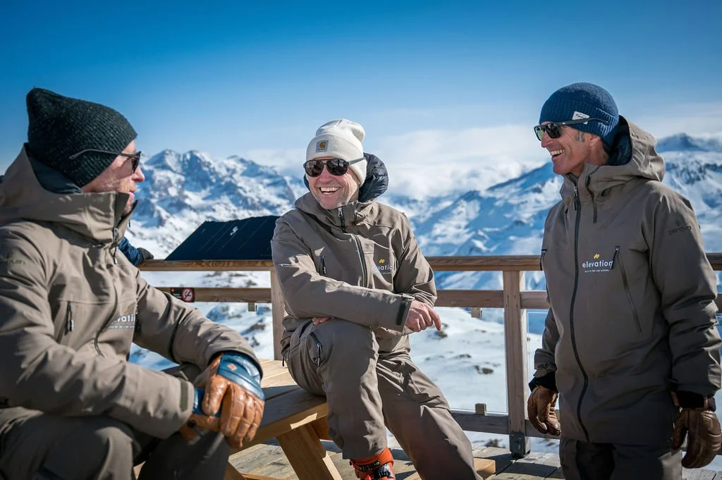 image shows 3 Elevation ski instructors in uniform in relaxed setting chatting and laughing with blue skies and mountains in background