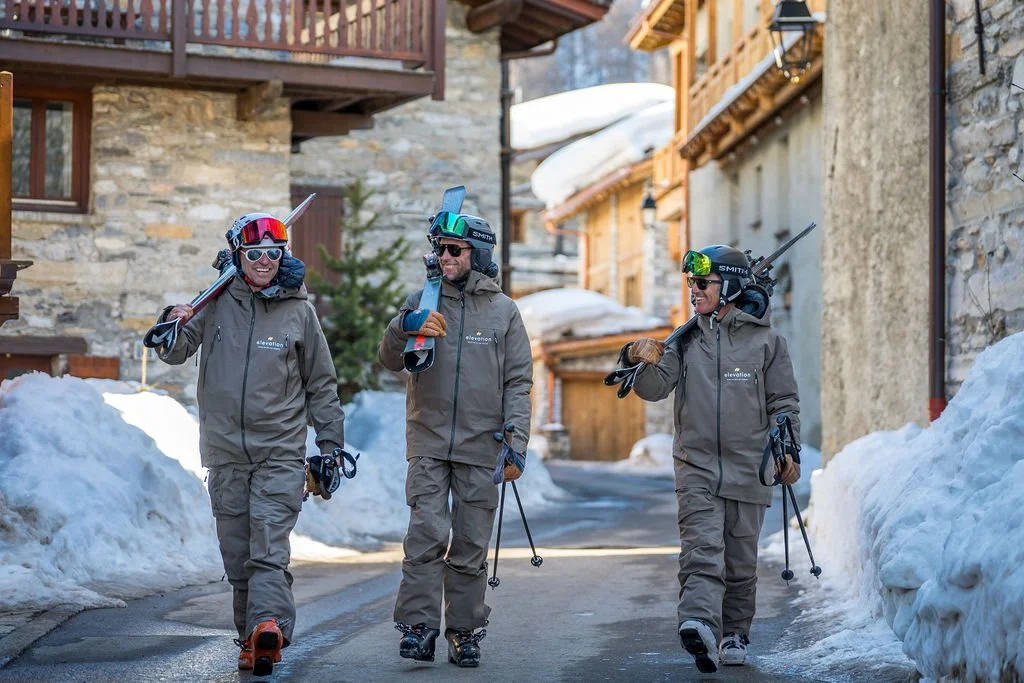 image show 3 Elevation ski instructors walking down street in old alpine village in uniform with skis on shoulders