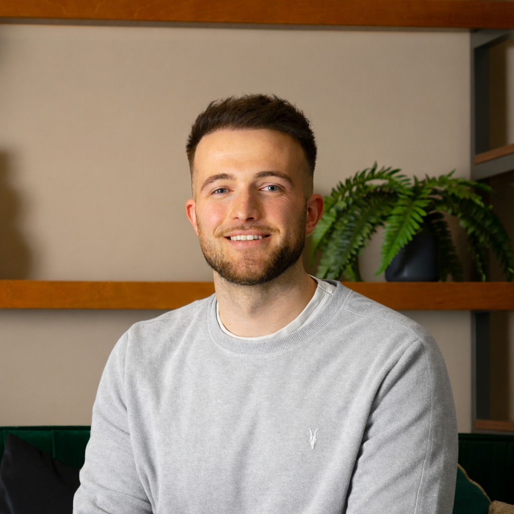 A smiling young man with short brown hair and a beard, wearing a light gray sweatshirt, sitting indoors in front of wooden shelves and a green plant.