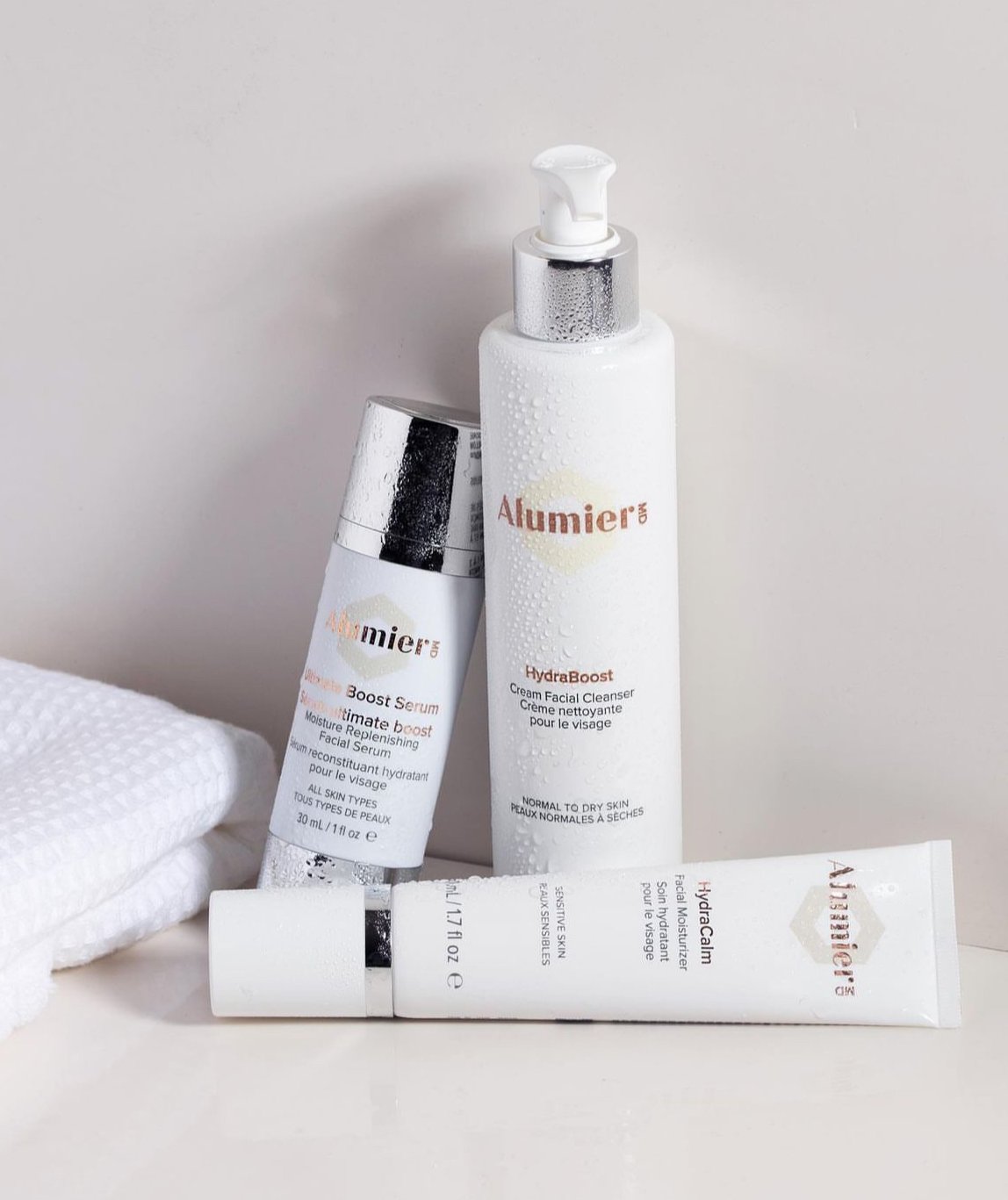 Three skincare products from Alumier MD, including a moisturiser, serum, and facial cleanser, along with a white towel on a white surface.