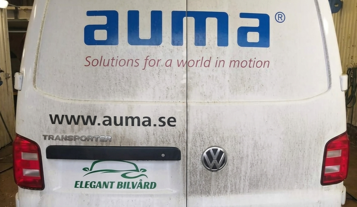 En ren, vit Volkswagen Transporter med reklamskyltar för Auma och Elegant Bilvård på bakluckan, bilen är smutsig och står i ett garage.