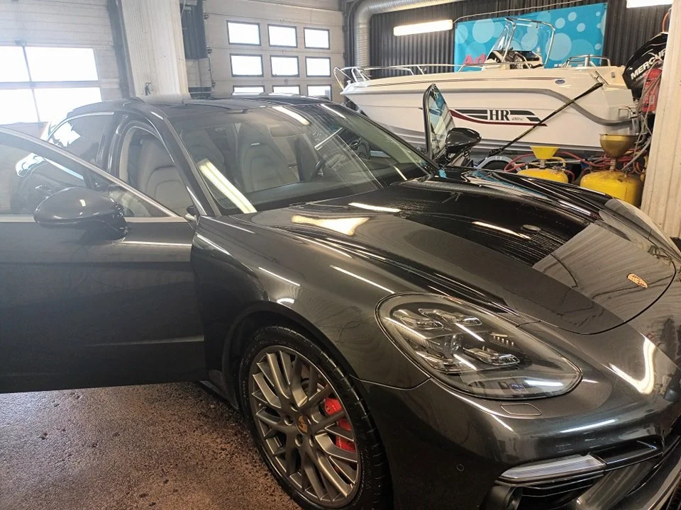En svart sportbil, troligen en Porsche, i ett garage. I bakgrunden syns en båt och några gula reservdelar.