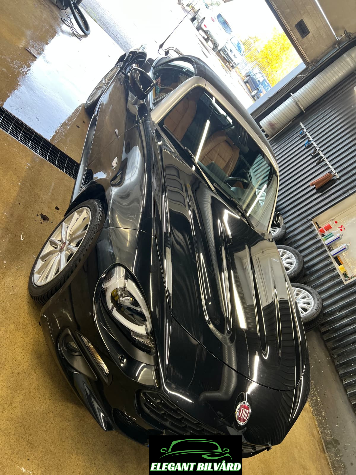 En svart Fiat bil är parkerad inomhus i en bilverkstad, med beige inredning i förarsätet. Verkstaden har hörnbelysning och på väggarna finns hyllor med rengöringsprodukter. Utomhus syns parkerade bilar och träd med höstfärgat löv.
