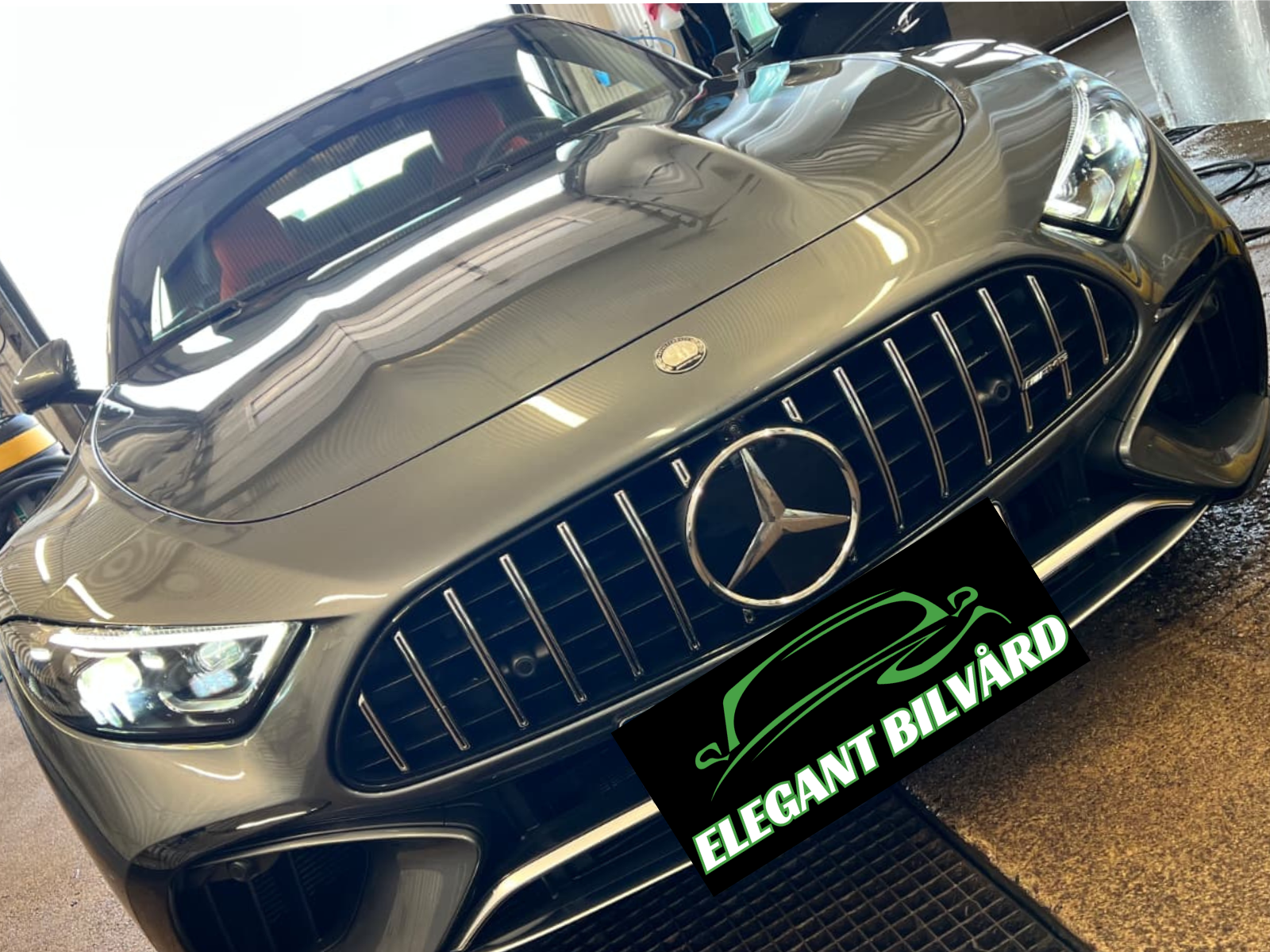 En silverfärgad Mercedes-Benz bil i ett garage, med en grön och vit skylt som säger 'ELEGANT BILVÅRD'.