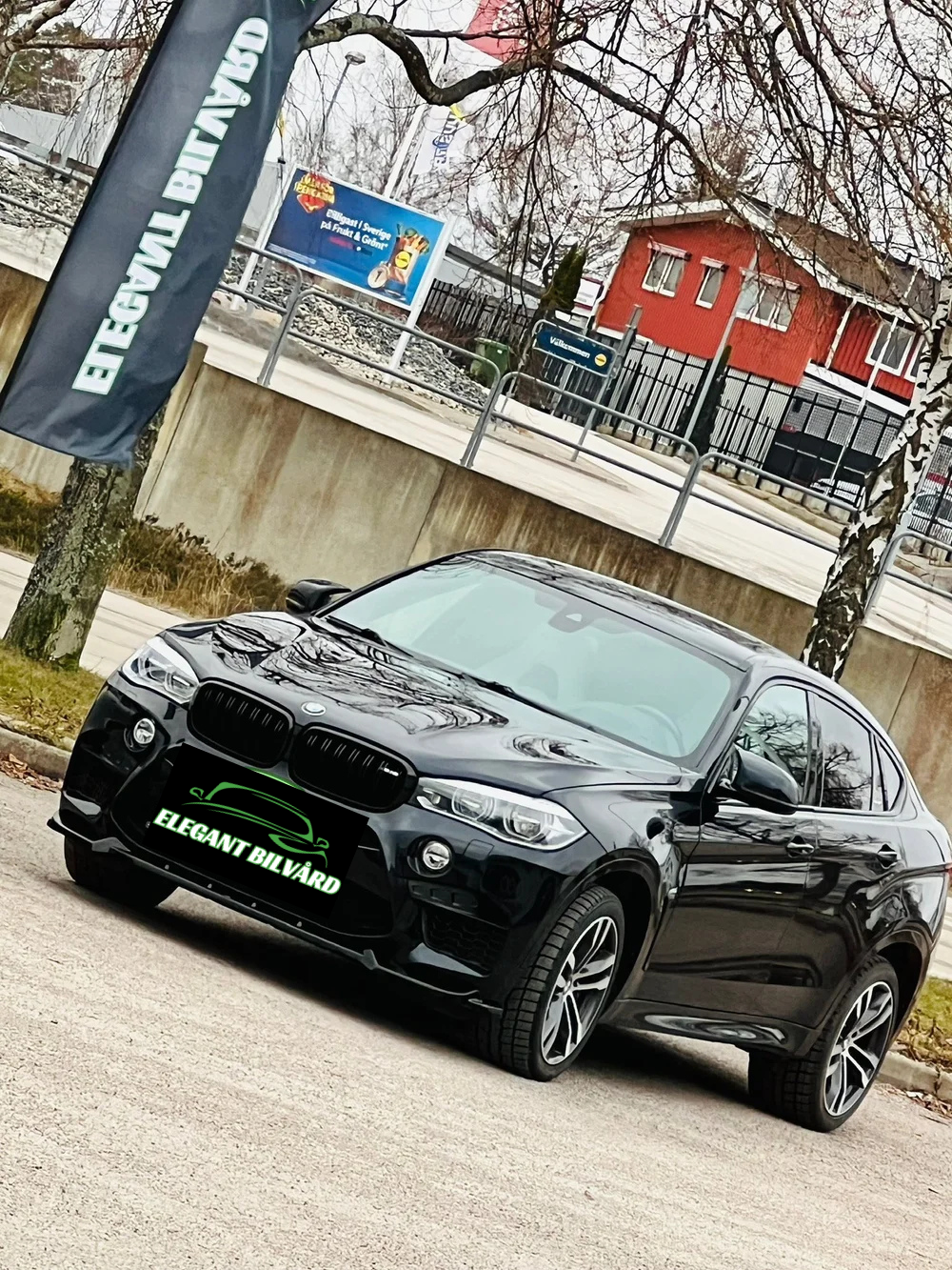 Svart BMW X4 bil parkerad nära trottoaren, med skylt på fronten som säger "ELEGANT BILVÅRD". Bakgrund med träd, en flagga, ett underlägg med texten "Välkommen," och ett rött hus.