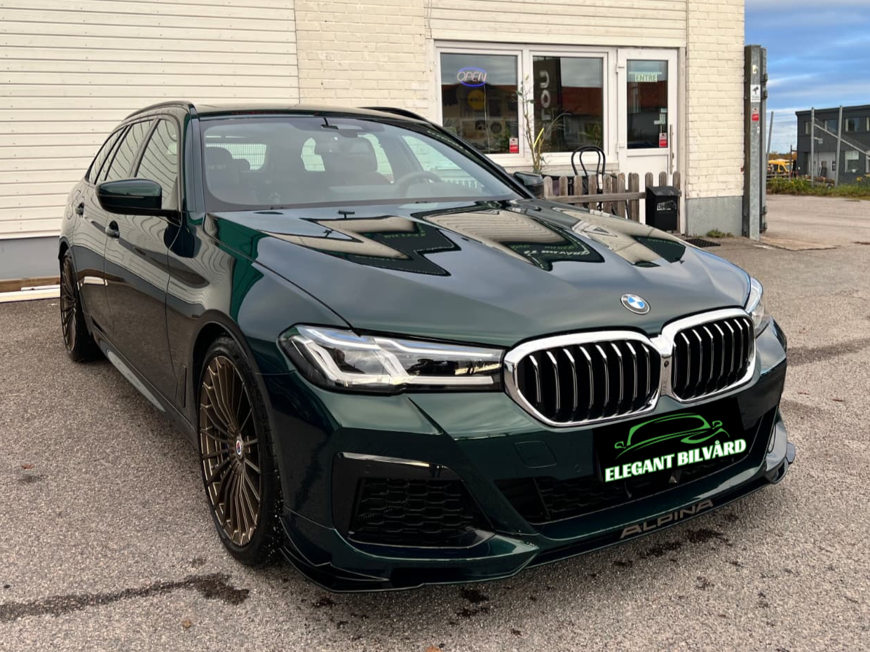 En mörkgrön BMW-camry parkerad utanför en vit byggnad med skyltar för 'Open' och 'Entré'.