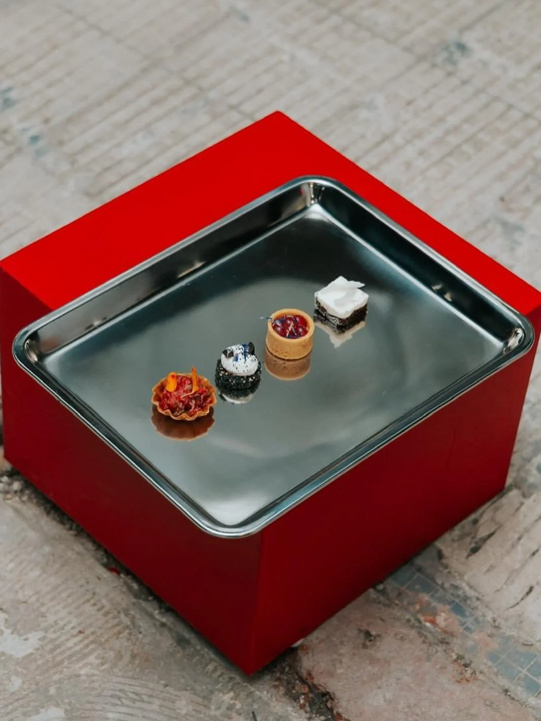 Quatre petites pâtisseries sur une plaque en métal, posée sur une surface rouge. La photo est prise en hauteur. Les pâtisseries semblent être des desserts sophistiqués.