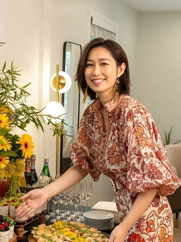 Woman smiling at a gathering, standing next to a table with snacks and flowers, wearing a patterned dress.