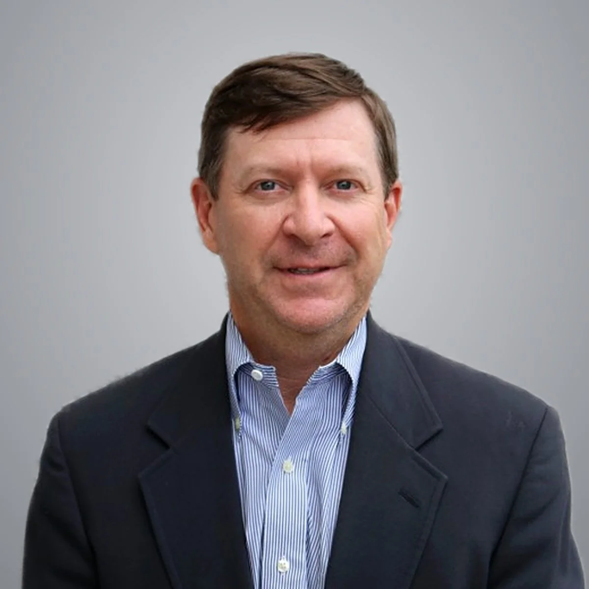 Headshot of a man with brown hair, blue eyes, wearing a dark blazer and a light blue striped shirt, against a plain gray background.